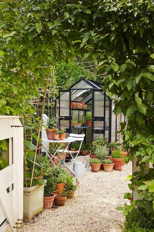 Small cottage greenhouse with black shelves in a gravel backyard finished with white bistro chairs and a table.