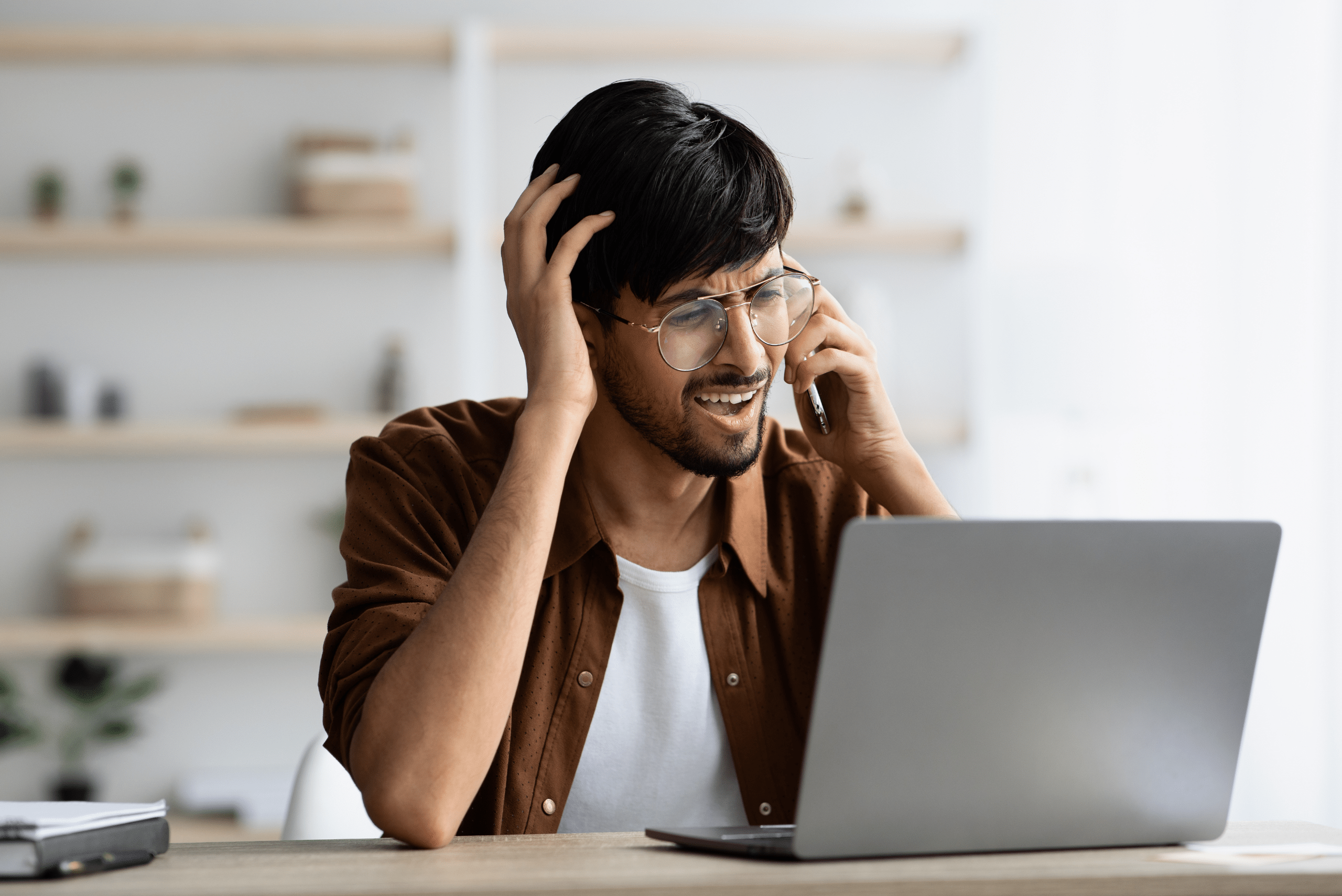 Man wearing glasses using a laptop and talking on the phone.