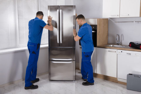 Two men in blue outfits moving a fridge.