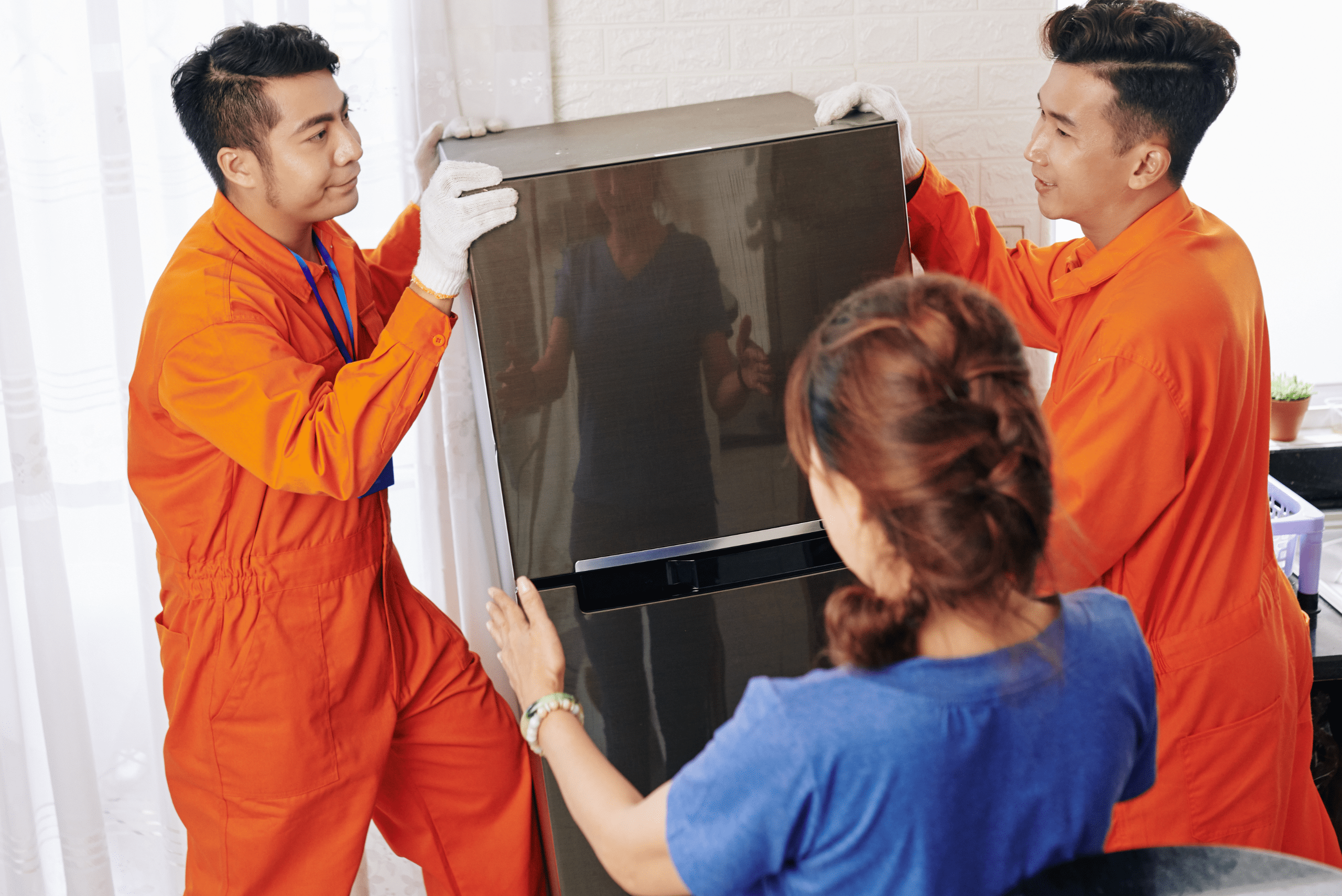 Two men in orange outfits moving a fridge with a woman directing them.